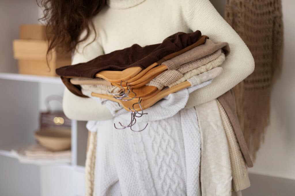 Une femme portant plusieurs pulls dans ses bras. Les pulls de différentes couleurs (noir, beige, marron, blanc) sont attachés à des ceintres avec des manches en bois.