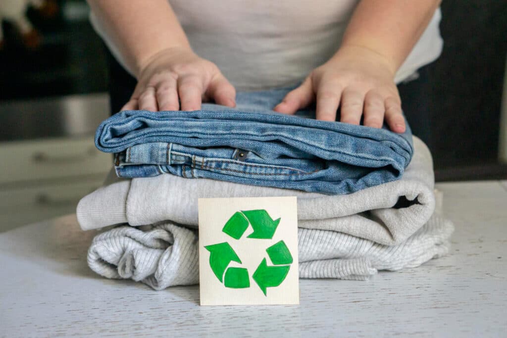 Une main au dessus d'un jean bleu et de 2 pulls de couleurs gris avec un petit carton sur lequel est inscrit le logo de recyclage de couleur verte.