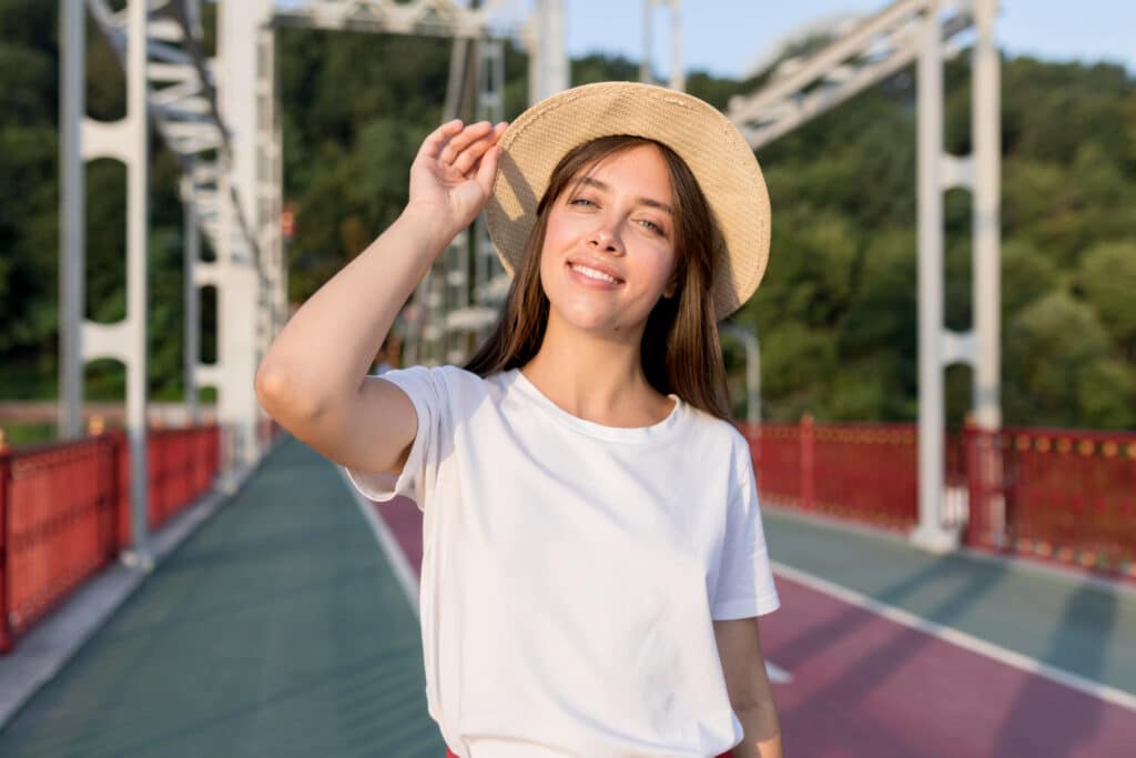 une belle jeune femme brune souriante qui porte un t shirt coton bio blanc uni et un chapeau. Elle se tient sur un pont avec un fond gris, violet, rouge et de la verdure.