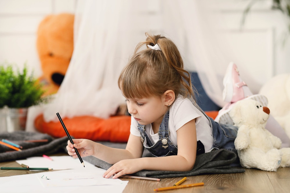 une petite fille avec un tshirt coton Oeko-tex et une salopette en jeans qui dessine sur du papier blanc, entourée de peluche dans sa chambre.