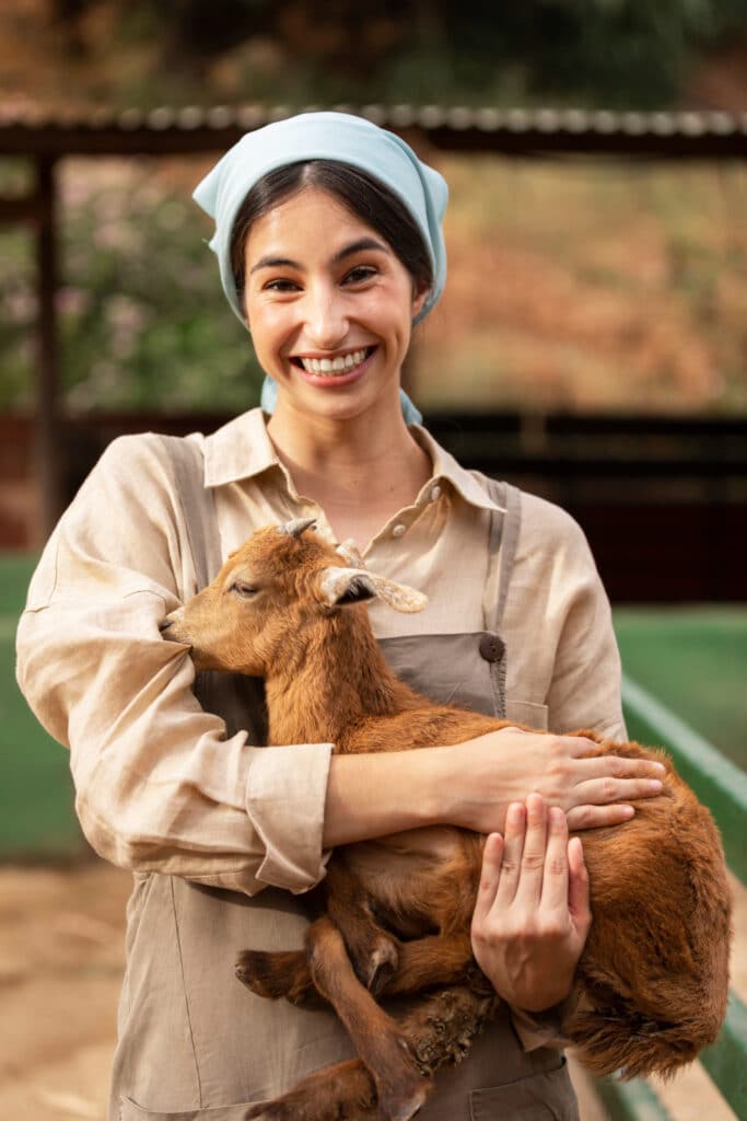 Une femme souriante portant un foulard bleu sur la tête, une chemise beige et une salopette marron, tenant dans ses bras, une petite chèvre de couleur marron.