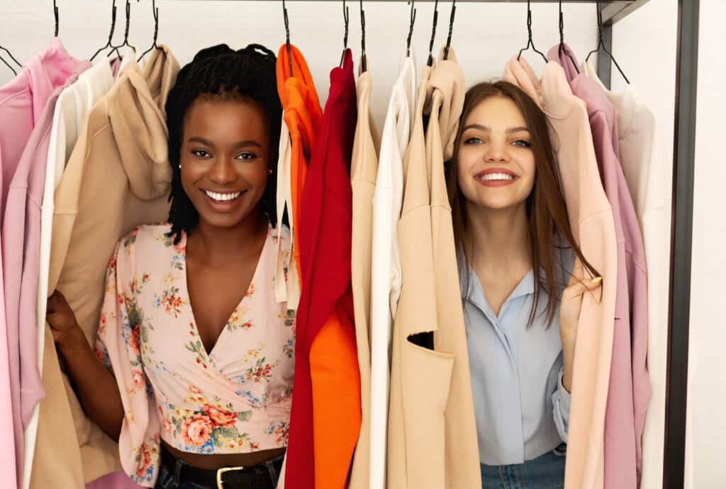 Deux jeunes femmes souriantes, l'une noire, l'autre blanche, se tenant entre des vêtements de différentes couleurs accrochés sur des cintres.