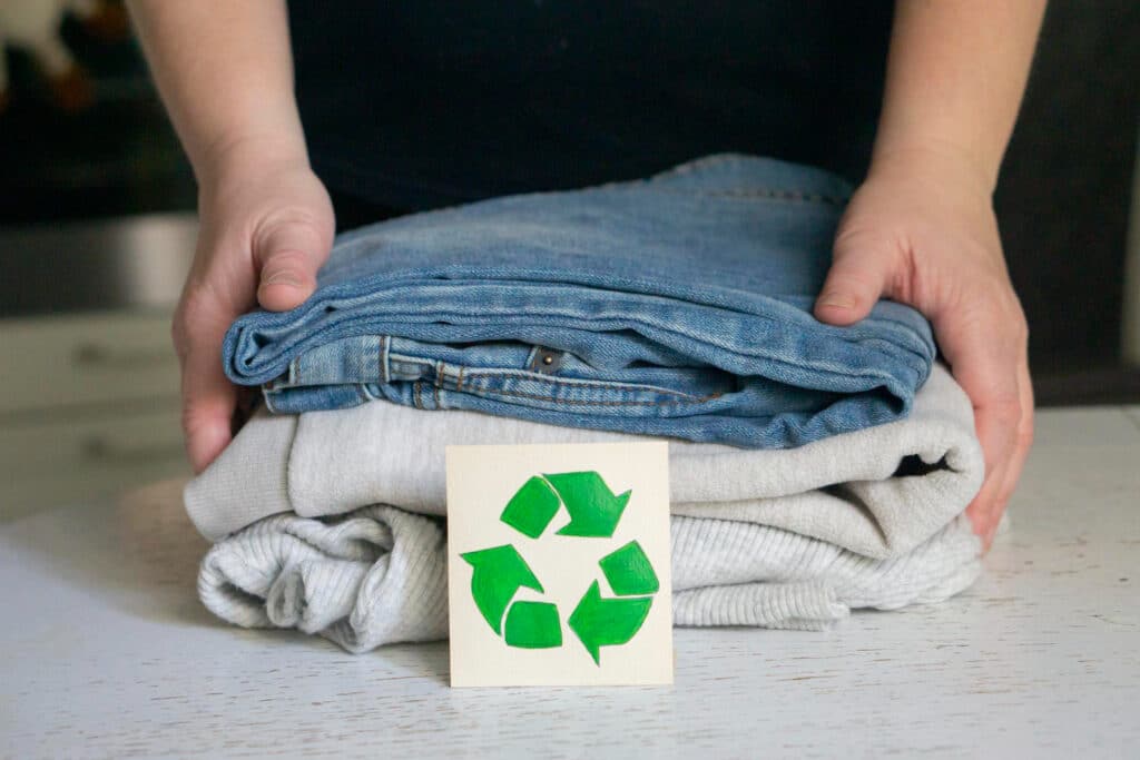 Une main tenant un jean bleu et un pull gris pliés. Devant se trouve un petit carton avec le logo de recyclage de couleur verte.