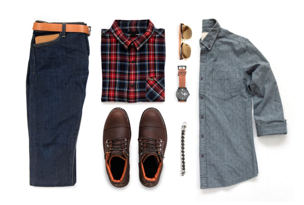 Des vêtements et accessoires pour homme: un jean bleu avec une ceinture marron, une chemise à carreaux rouge et bleu, des chaussures en cuir marron, une montre, une paire de lunette, un bracelet et un chemise gris à motif.