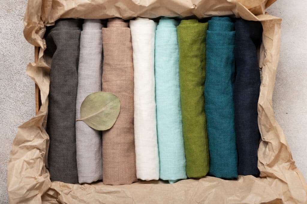 Des tissus en lin de différentes couleurs: bleu marine, cyan, vert mousse, bleu clair, beige et gis, pliés et rangés dans un emballage en papier.